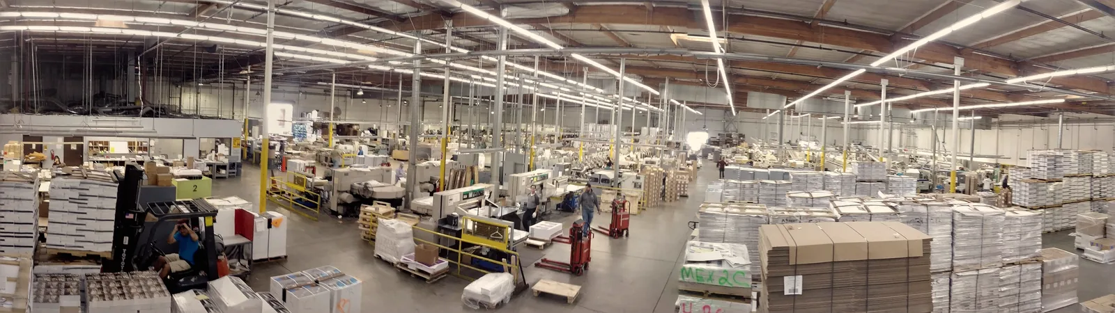 Wide view of the Ross Bindery production floor: rows of binding and finishing equipment under high bay lighting, with pallets of printed sheets and stacked cartons staged along the aisles.